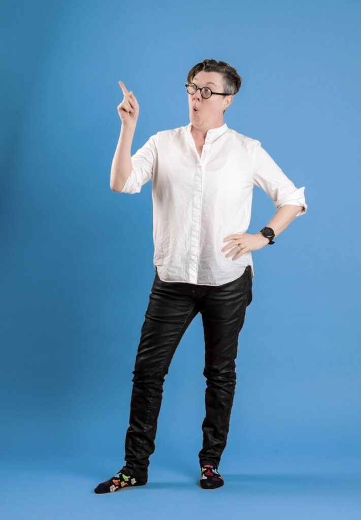 Person standing against a bright blue background wearing a white shirt, black trousers and colourful socks, pointing upwards with an animated expression.